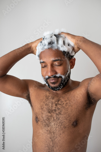 Young handsome smiling African American man with a beard washing his hair with foamy anti-dandruff shampoo in a bathroom, his hair covered in foam