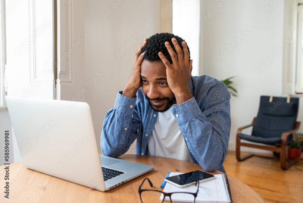 Frustrated young African entrepreneur with sad grimace in front of his laptop at home office, noticed a big mistake he did, holding his face with hands, wearing blue shirt Frustrated young African entrepreneur with sad grimace in front of his laptop at home office, noticed a big mistake he did, holding his face with hands, wearing blue shirt
