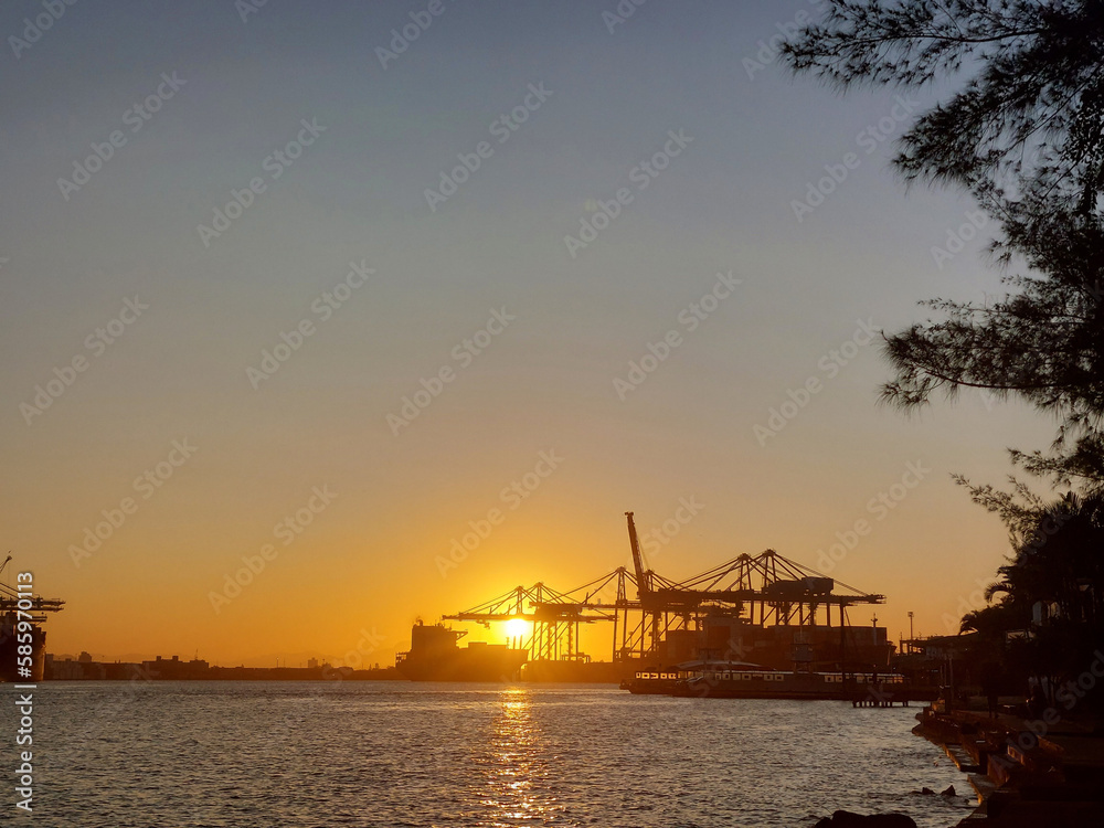 Behind the silhouette of trees and a commercial port, we can see the bright golden sun setting on the horizon, coloring the sky in shades of orange, reaching blue and illuminating the calm waters of a