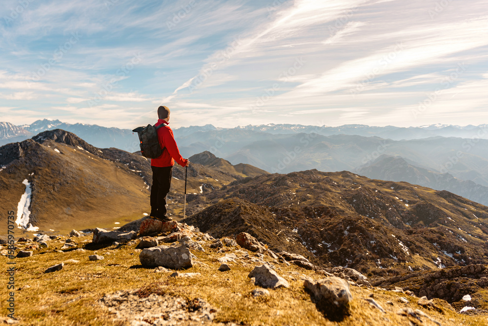 Obraz premium mountaineer man from behind with red clothes, backpack and trekking poles practicing high mountain hiking and contemplating the landscape. outdoor sport and adventure.