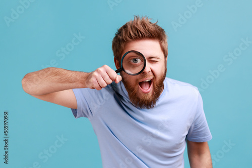 Man in blue T-shirt holding magnifying glass and looking at camera with big zoom eye, curious face.