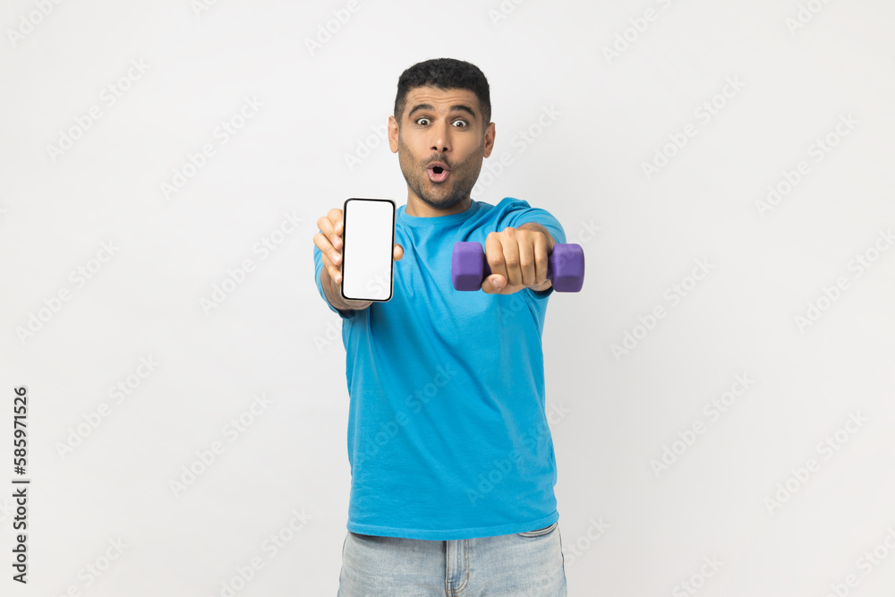 Shocked man holds dumbbell and smartphone with empty display advertisement for fitness app.