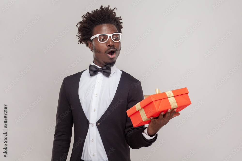 Surprised astonished man in glasses standing with red gift box, looking at present with open mouth.