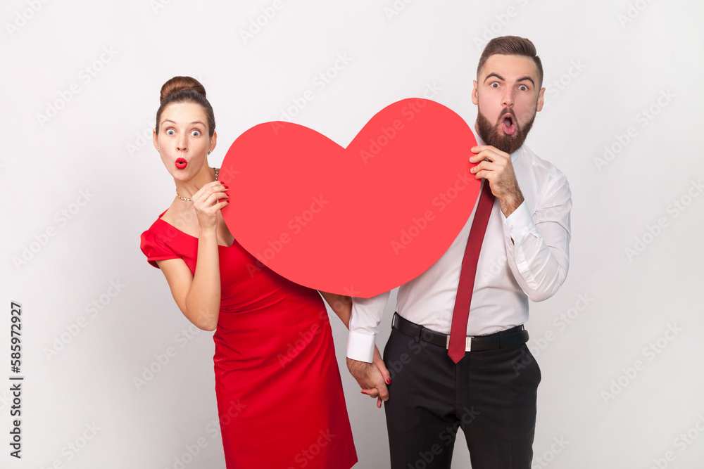 Amazed shocked man in white shirt, woman in red dress standing together, peeping out from big heart.
