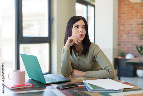 pretty young woman smiling with a happy, confident expression with hand on chin. laptop and desk concept