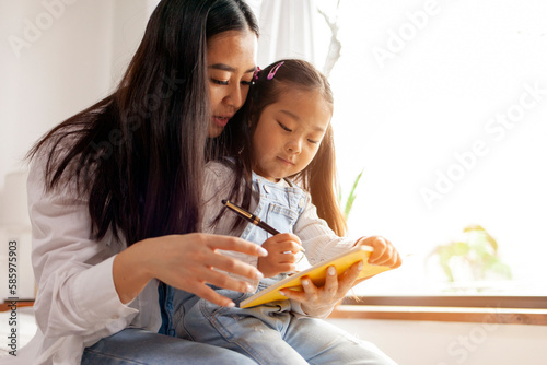 asian woman teaches her little daughter to write and draw in notebook at home, korean girl with her mother and write