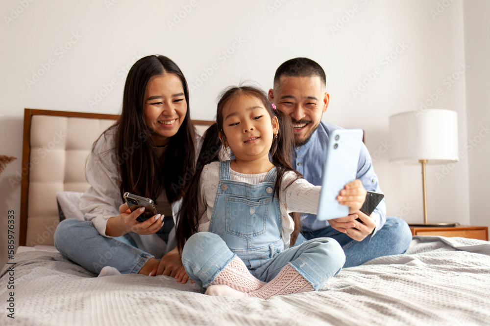 asian family sit on the bed and communicate via video call, little korean girl with mom and dad takes selfie asian family sit on the bed and communicate via video call, little korean girl with mom and dad takes selfie