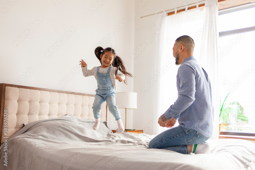 small active Asian girl plays with her dad at home, energetic child jumps and dances on the bed with parent small active Asian girl plays with her dad at home, energetic child jumps and dances on the bed with parent