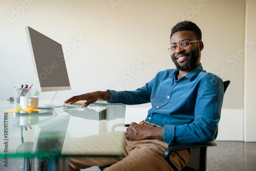 Portrait of positive african american male entrepreneur sitting in front of computer, smiling at camera, free space