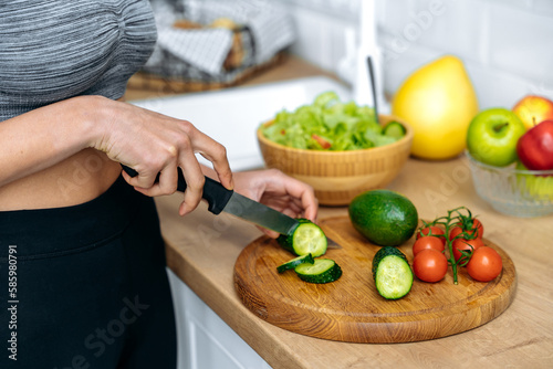Healthy eating concept. Close-up hands of african american young woman in a sportswear, standing at home in kitchen cutting cucumber while preparing fresh useful salad, smiling happily