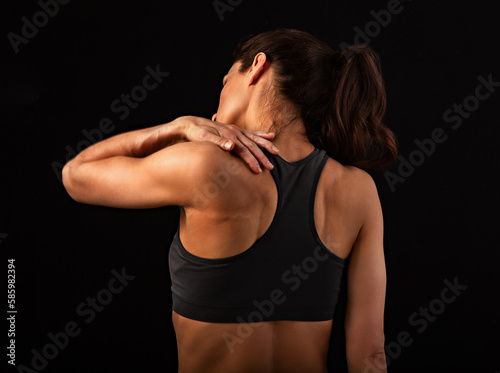 Female sporty muscular with ponytail doing stretching workout of the shoulders, blades in sport bra, holing the neck the hand on black studio background © nastia1983