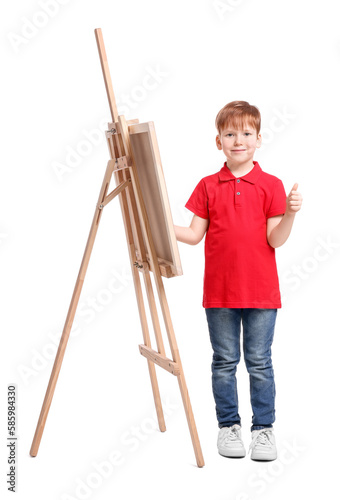 Little boy with brush near easel with canvas showing thumbs up against white background. Creative hobby