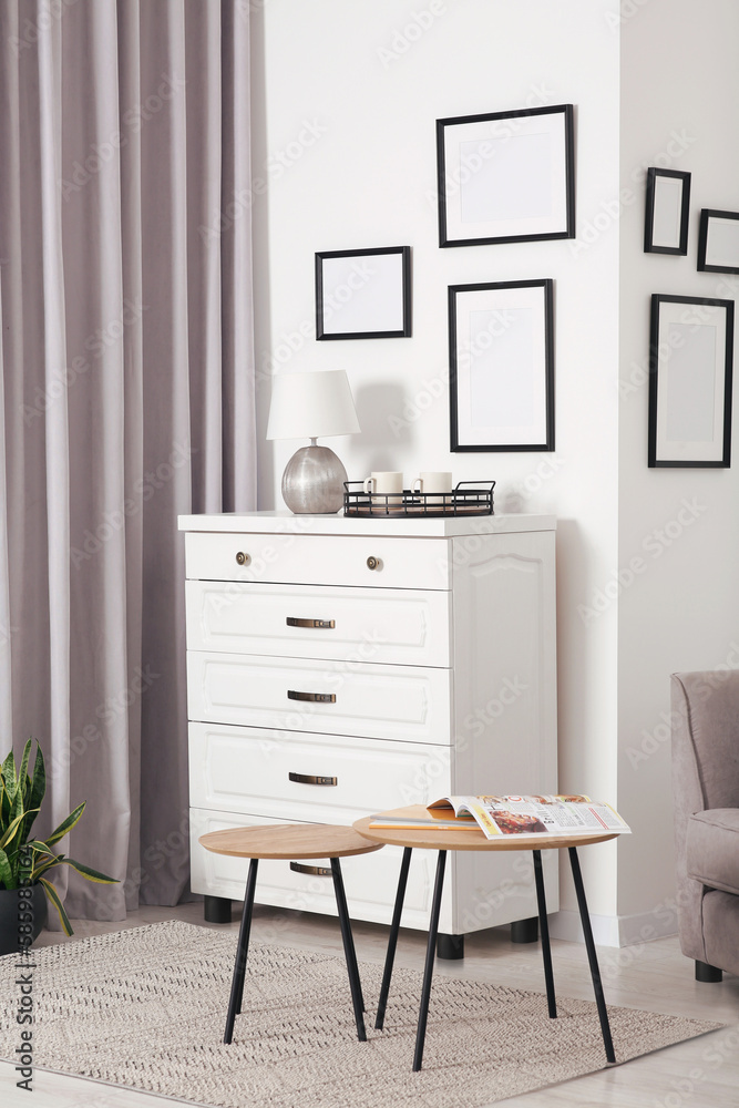 Fototapeta premium Empty frames hanging on white wall, chest of drawers and wooden tables indoors