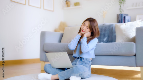 Office syndrome concept. Asian young woman working on laptop while seated on floor next to couch at home, feeling pain in neck and shoulder after working on computer laptop for a long time