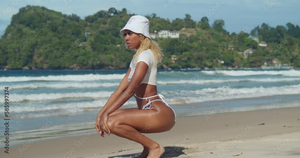 Bikini girl squats in the sand of a tropical island beach with ocean waves in the background Bikini girl squats in the sand of a tropical island beach with ocean waves in the background