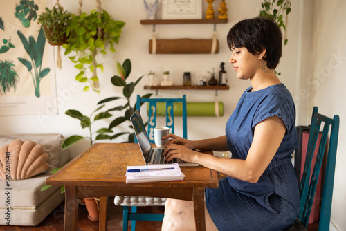 Young latin woman with short hair wearing a blue dress. She is sitting in the apartment full of plants and using a laptop.