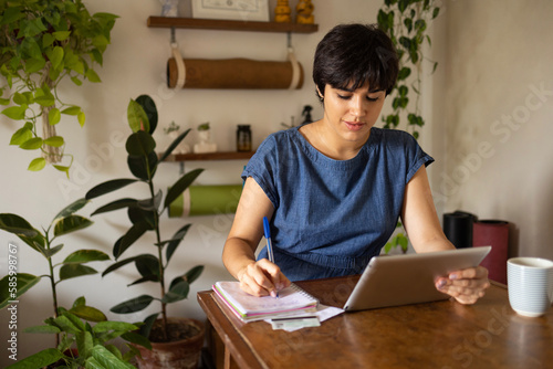 Young latin woman with a short hair wearing a blue shirt and taking notes from a tablet at the home office full of green plants