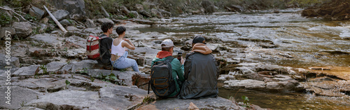 Rear on two couples of tourists sitting and resting at rocky river valley in mountains.