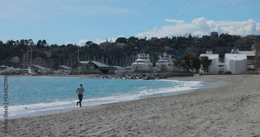 Male Seen Running Along Beach Shoreline At Varazze, Italy On Sunny Clear Day. Locked Off Male Seen Running Along Beach Shoreline At Varazze, Italy On Sunny Clear Day. Locked Off