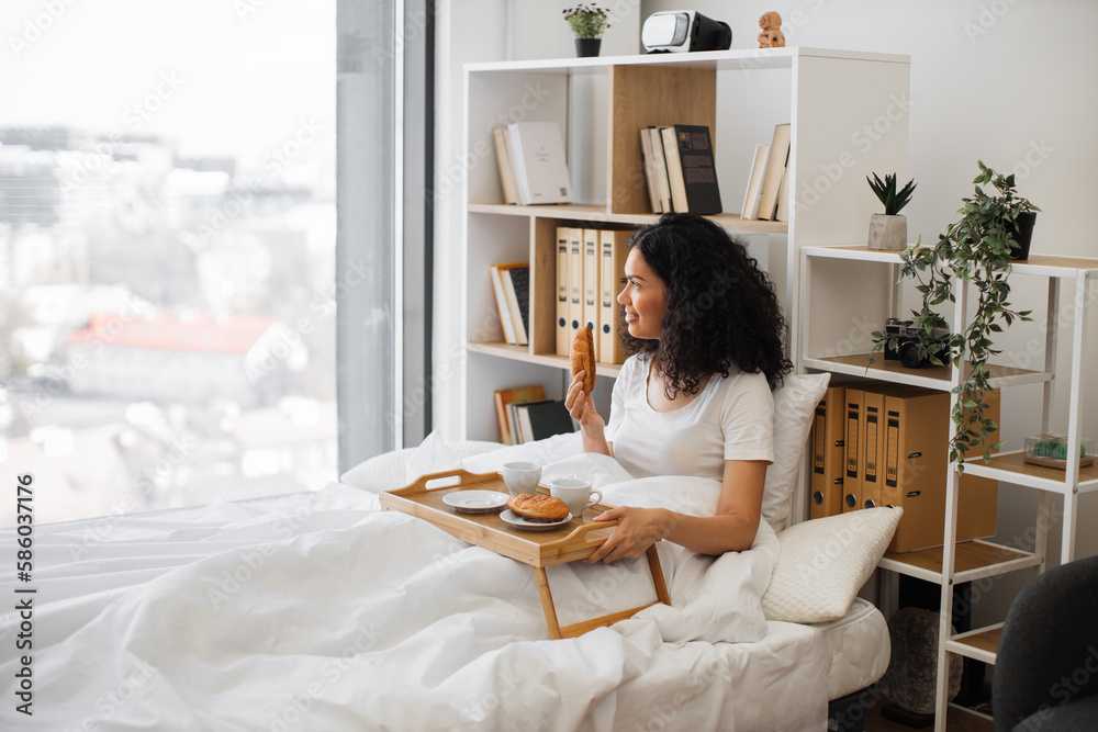 Beautiful young lady holding delicious croissant in hand while enjoying view out of panoramic window of white bedroom in morning. Pretty woman waiting for her husband to join for coffee in bed. Beautiful young lady holding delicious croissant in hand while enjoying view out of panoramic window of white bedroom in morning. Pretty woman waiting for her husband to join for coffee in bed.