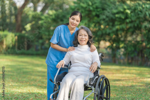 Young asian care helper with asia elderly woman on wheelchair relax together park outdoors to help and encourage and rest your mind with green nature. Looking camera. © NanSan