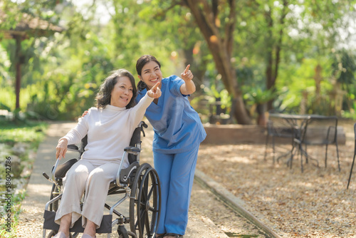 Young asian care helper with asia elderly woman on wheelchair relax together park outdoors to help and encourage and rest your mind with green nature. Pointing