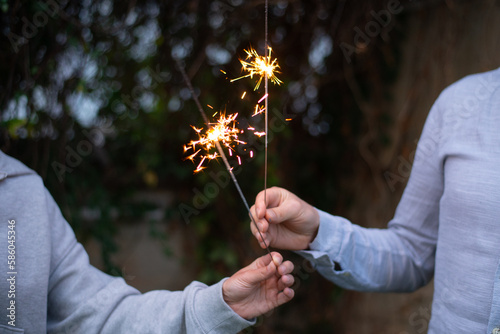 Female and male hands holding sparkling fireworks at twilight against trees in the evening. Couple celebrating holiday