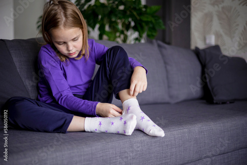 A cute little blonde girl puts white socks with stars on her feet. Independence of children. The development of dress skills in younger schoolchildren.