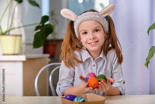 A positive playful girl in bunny ears holds colorful eggs in her hands in the home kitchen. Happy Easter. Easter background