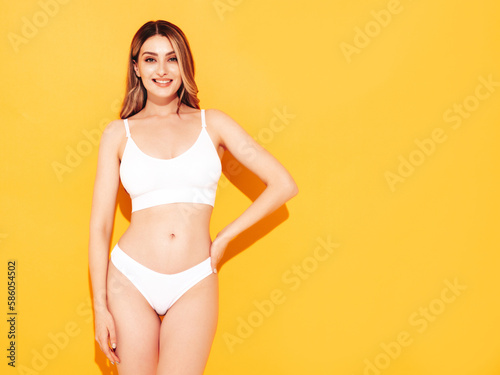 Fashion portrait of young beautiful woman. Attractive carefree blond model wearing pure white lingerie. Hot tanned smiling blonde posing near yellow wall in studio, showing sexy natural body