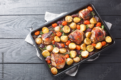 Schilderij op canvas Chicken thighs baked with potatoes, garlic, onions and tomatoes close-up on a black baking sheet on a wooden table