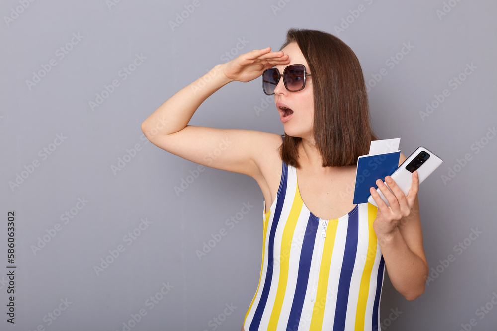 Indoor shot of amazed surprised woman looking far with hand on forehead at copy space for promotion tourist holding cellphone and passport wearing striped swimming suit isolated over gray background