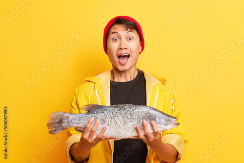 Studio shot of Asian fisherman standing over yellow background and holding a fish with happy expression emotions dressed in red hat and yellow fishing jacket. People emotions concept