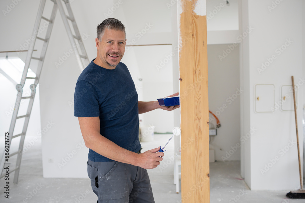 Middle-aged man with grey hair painting wooden beams in attic apartment, construction site with white paint Middle-aged man with grey hair painting wooden beams in attic apartment, construction site with white paint