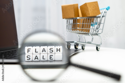 Flash sale word on cubes through a magnifying glass and with a toy shopping cart with boxes on the background. Online shopping concept. 