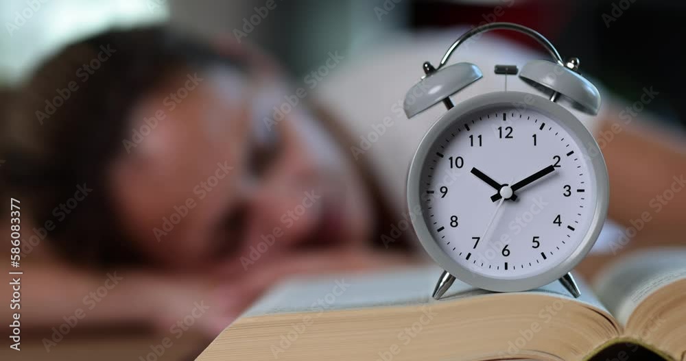 Alarm clock stands on open book against sleeping woman