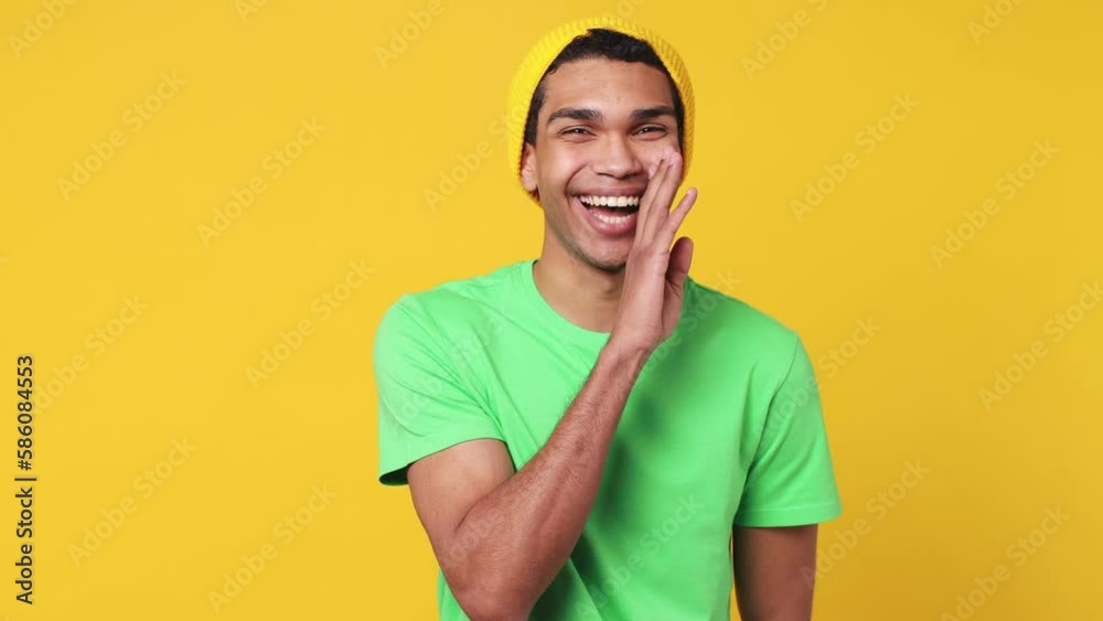 Mysterious joyful young man of African American ethnicity wears green t-shirt hat whispering gossip and tells secret behind his hand sharing news isolated on plain yellow background studio portrait Mysterious joyful young man of African American ethnicity wears green t-shirt hat whispering gossip and tells secret behind his hand sharing news isolated on plain yellow background studio portrait