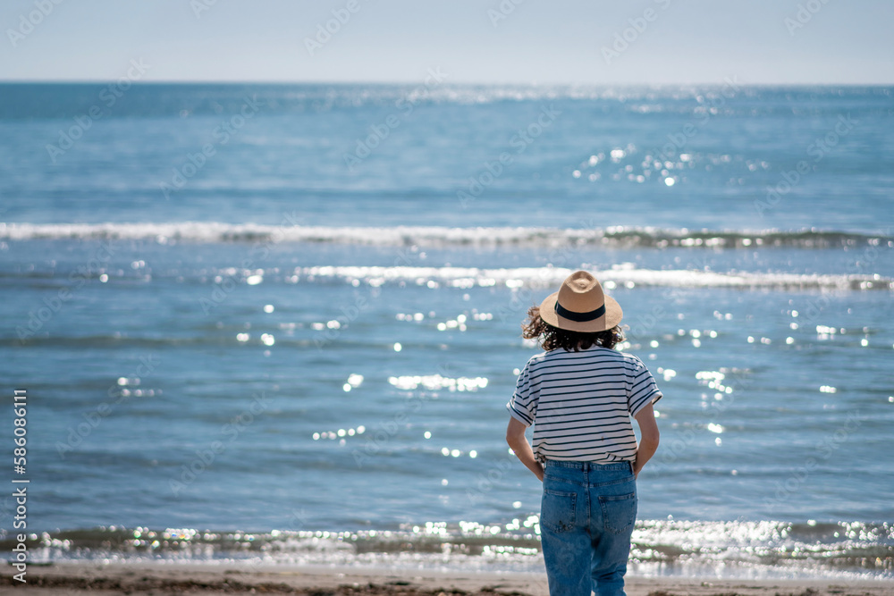 Young woman in a hat and a striped t-shirt walking towards the blue sea on a summer day