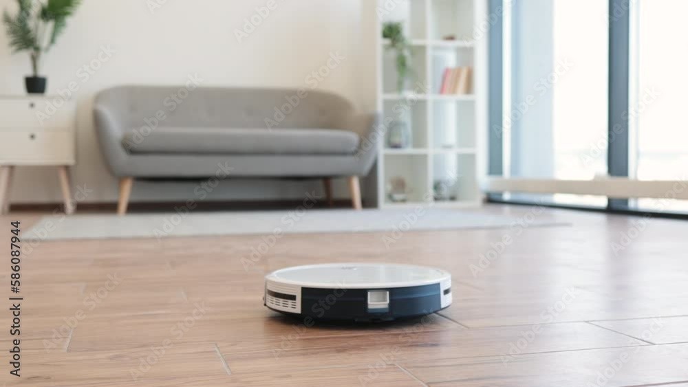 Automatic robot vacuum moving from soft carpet to hardwood floor while removing dust in living room. Smart home device conducting daily vacuuming and saving residents from seasonal allergies. Automatic robot vacuum moving from soft carpet to hardwood floor while removing dust in living room. Smart home device conducting daily vacuuming and saving residents from seasonal allergies.