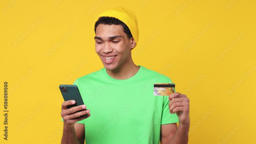 Young man of African American ethnicity wear green t-shirt hat use mobile cell phone app hold credit bank card do online shopping order delivery to home look aside isolated on plain yellow background Young man of African American ethnicity wear green t-shirt hat use mobile cell phone app hold credit bank card do online shopping order delivery to home look aside isolated on plain yellow background