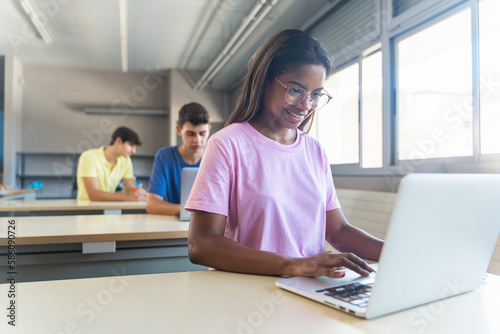 Smiling African American female student wearing eyeglasses standing using laptop in classroom