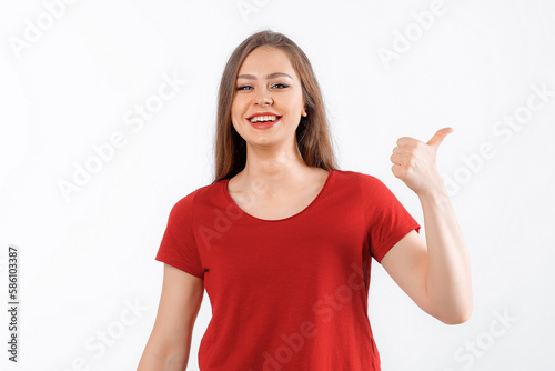 Smiling lovely girl points finger at promo banner, shows advertisement on blank white space. Young brunette female looking happy by promotion offer, stands over white background