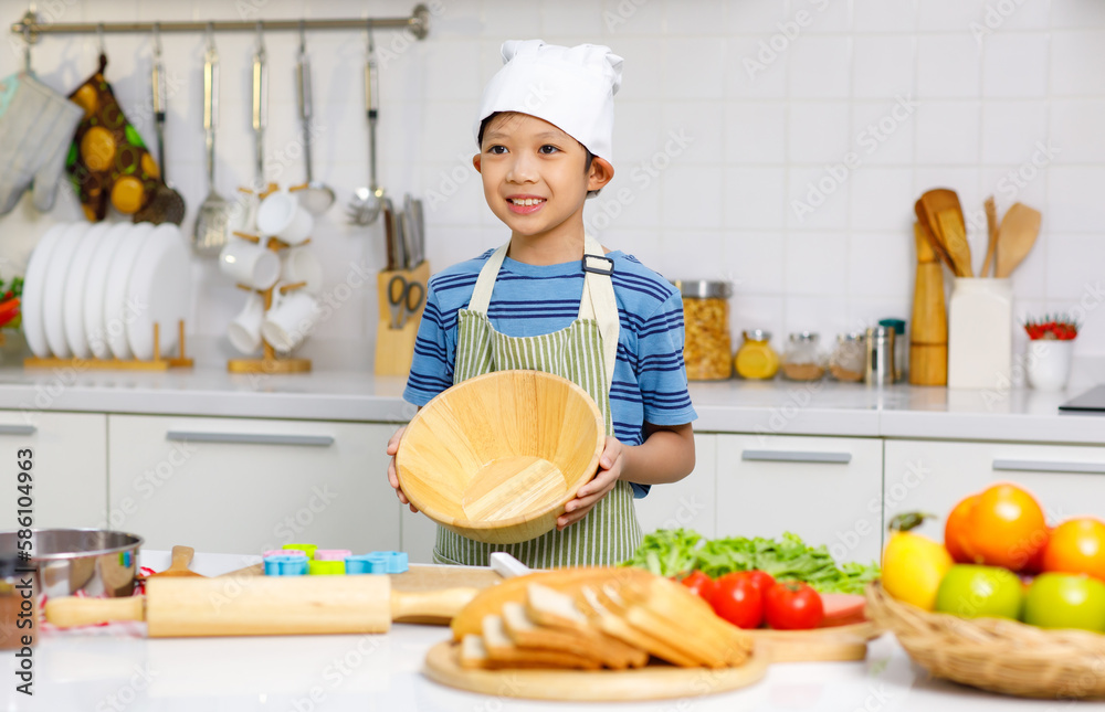 Millennial Asian young little boy chef wearing white tall cook hat and apron standing smiling holding wooden bowl posing taking photo while preparing cooking fresh meal at counter in home kitchen Millennial Asian young little boy chef wearing white tall cook hat and apron standing smiling holding wooden bowl posing taking photo while preparing cooking fresh meal at counter in home kitchen
