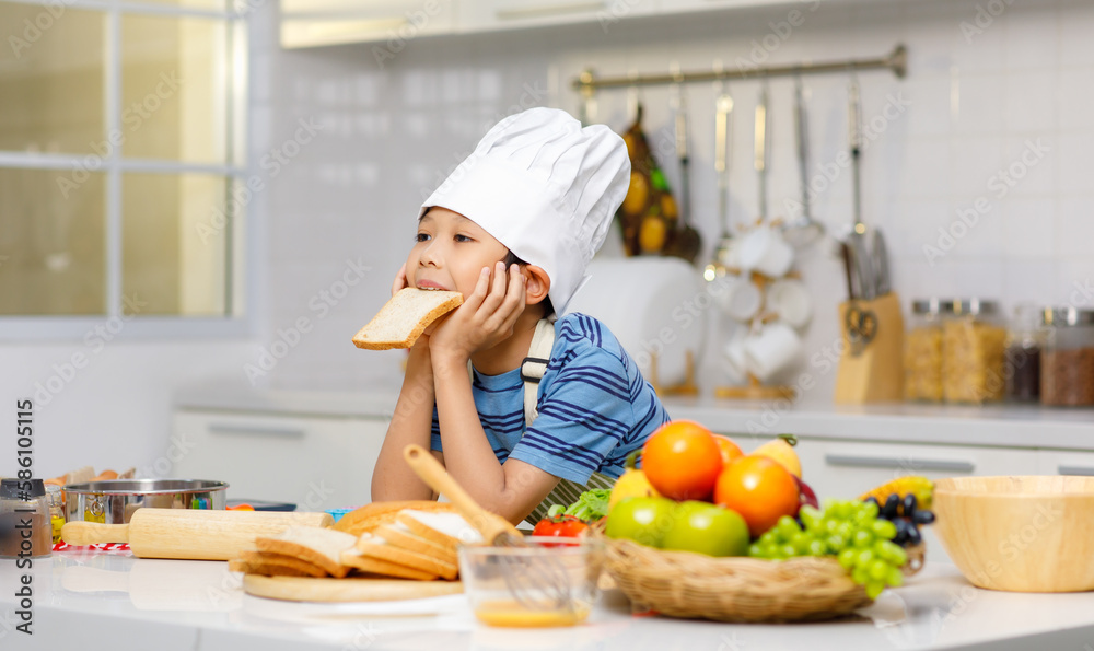Millennial Asian little boy chef wearing tall white cook hat and apron standing posing eating sliced bread in home white kitchen full of fruits vegetables and bread on cooking counter. Millennial Asian little boy chef wearing tall white cook hat and apron standing posing eating sliced bread in home white kitchen full of fruits vegetables and bread on cooking counter.