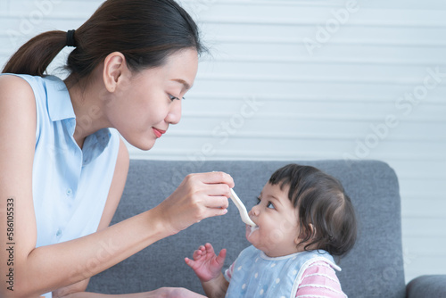 Selective focus on young mother hand is holding spoon feeding Caucasian Cute 7 month newborn baby girl  child eating blend mashed food  sitting on sofa with apron at home  baby mouth mess up with food