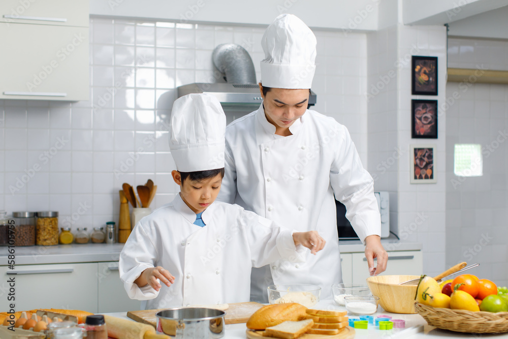 Asian young little boy chef in uniform with white tall cook hat standing learning preparing massaging flour dough on wooden cutting board while male cooking teacher helping teaching in home kitchen