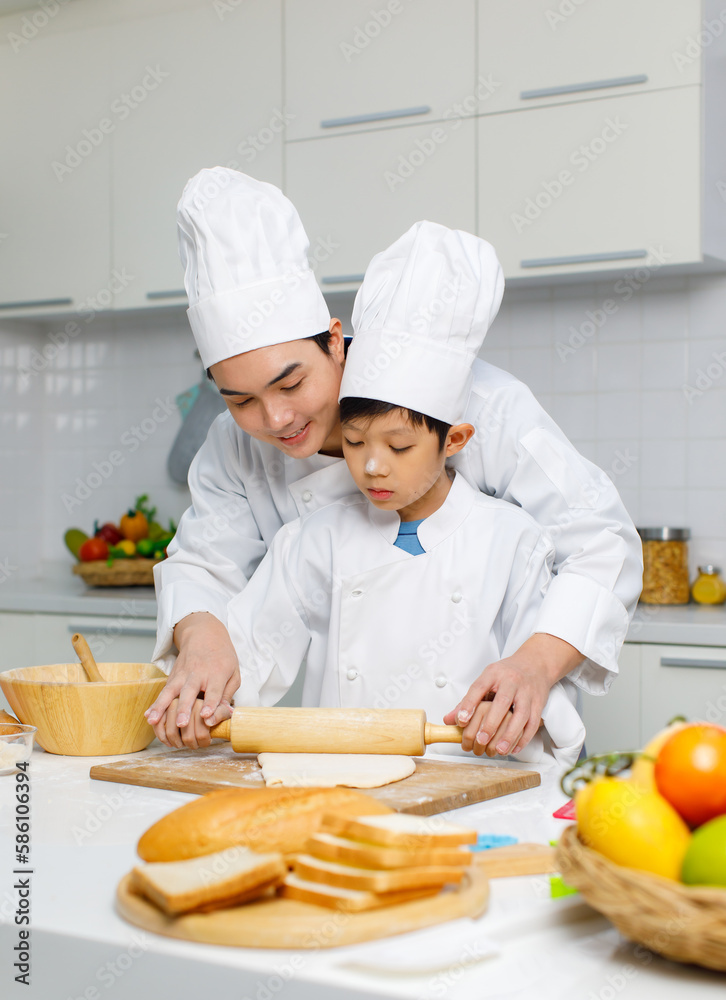Asian young little boy chef in uniform with white tall cook hat standing learning preparing massaging flour dough on wooden cutting board while male cooking teacher helping teaching in home kitchen Asian young little boy chef in uniform with white tall cook hat standing learning preparing massaging flour dough on wooden cutting board while male cooking teacher helping teaching in home kitchen