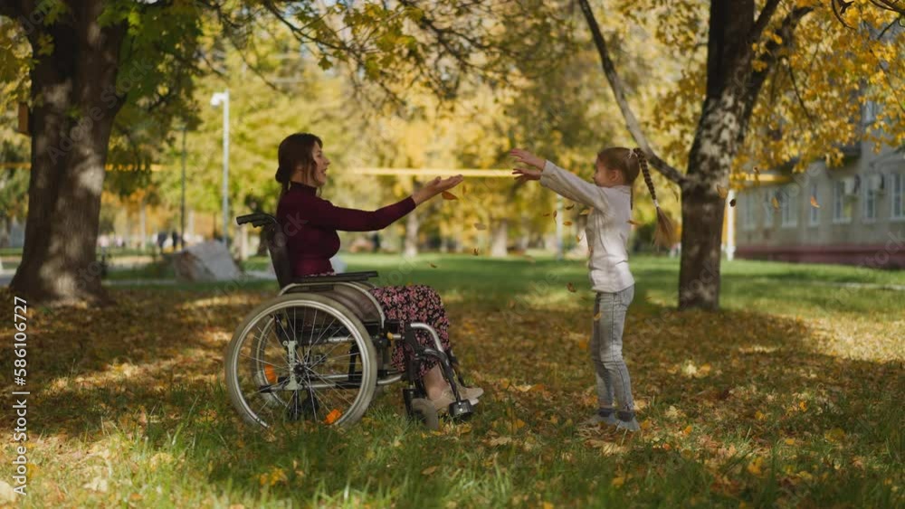 Positive adult woman with spinal cord injury in wheelchair and preschooler girl play throwing autumn leaves upward. Active family recreation during rehabilitation Positive adult woman with spinal cord injury in wheelchair and preschooler girl play throwing autumn leaves upward. Active family recreation during rehabilitation