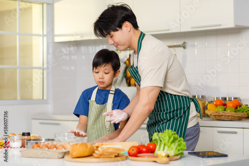 Asian young little boy chef wearing apron standing using stainless filter sifting white flour into glass bowl while father helping at counter full of baking equipment eggs breads tomatoes in kitchen