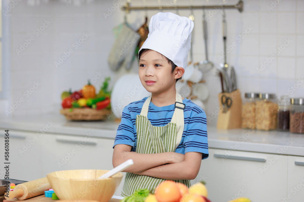 Millennial Asian little boy chef wearing tall white cook hat and apron standing posing in home white kitchen full of fruits vegetables and bread on cooking counter. Millennial Asian little boy chef wearing tall white cook hat and apron standing posing in home white kitchen full of fruits vegetables and bread on cooking counter.